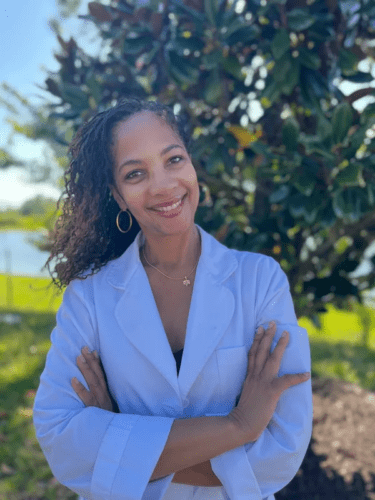 Smiling woman in a light blue blazer outdoors with greenery background.